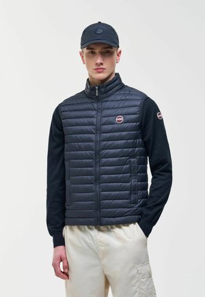 Giovane uomo in piedi di fronte, indossa un piumino blu navy trapuntato, maglia a maniche lunghe nera, berretto abbinato e pantaloni cargo beige con le mani nelle tasche.