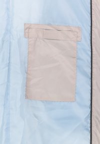 Jasnoniebieska tkanina z beżową prostokątną kieszenią naszywaną, z klapką u góry, na jasniejszym beżowym tle.