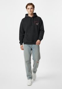 Felpa nera con cappuccio, con texture a coste, con un design di un cuore rosso e doppie rose sul petto, abbinata a jeans chiari strappati e sneakers bianche.