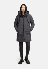 Grijze gewatteerde parka met een rits aan de voorkant, zijzakken en trekkoorddetails, gecombineerd met zwarte strakke pantalons en zwarte enkellaarsjes.