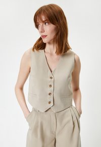 Gilet sans manches beige avec un décolleté en V profond, fermeture avant à cinq boutons et deux poches latérales. Coupe ajustée et tissu léger.