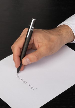 Roka drži črno pero in piše ročno pismo na belem papirju na črni površini.