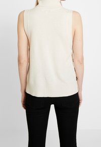 Beige ärmlös tröja med hög hals, med ribbad textur och sidoknappar, kombinerad med svarta slim fit-jeans.