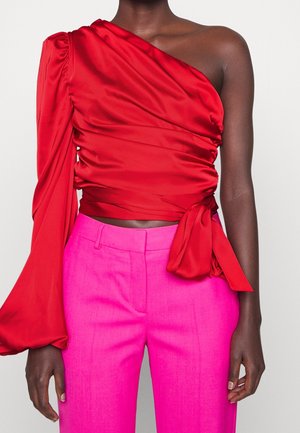 Osoba o ciemnej karnacji ubrana w jaskrawoczerwoną bluzkę na jedno ramię z długim rękawem, związaną w talii, oraz żywiołowo różowe, wysokowodne spodnie.