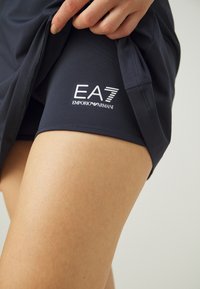 Pantalones cortos deportivos azul marino hechos de una tela suave, con el logo de EA7 Emporio Armani en blanco en la parte inferior de la pierna. Cintura elástica visible.