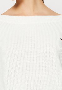 Maglione bianco a maglia testurizzata con design off-shoulder e piccolo ricamo. Presenta materiale a coste e una vestibilità larga.