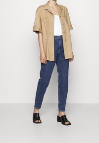 Ljusbeige kortärmad skjorta, vit linne, mörkblå tajta jeans och svarta klackar. Släta texturer och enkel design.