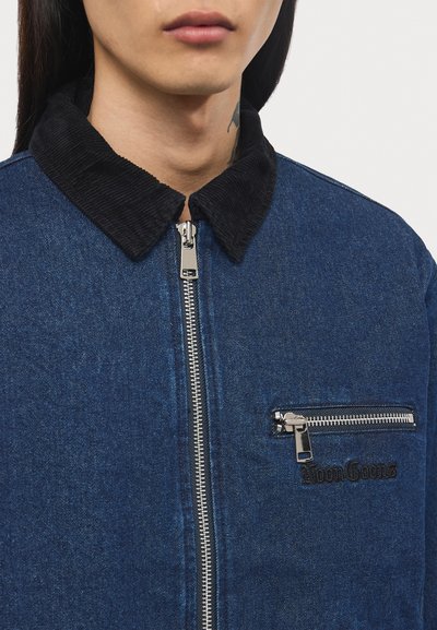 Denim jacket featuring a black corduroy collar, silver zipper, and a chest pocket with a zip closure and logo. Dark blue color.