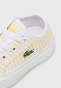 Baskets en toile blanches avec des rayures jaunes et blanches, arborant un logo Lacoste vert, bout rond et lacets blancs. Semelle en caoutchouc texturée.