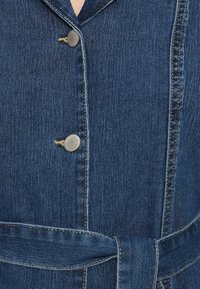 Τζιν φόρεμα με γιακά, κλείσιμο με ασημένια κουμπιά και ζωνάκι στη μέση. Το ύφασμα είναι σκούρο μπλε με διακριτή υφή και ραφές αντίθεσης.