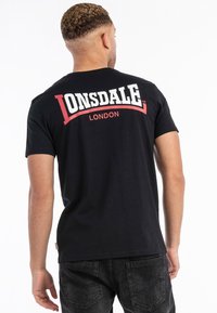 Mustahuivi puuvillapaita, jossa on valkoinen ja punainen "LONSDALE LONDON" -logosuunnittelu yläselässä. Lyhyet hihat ja klassinen istuvuus.