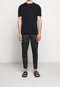 Marinblå bomullströja med korta ärmar, ihop med grå tapered byxor och svarta slip-on sandaler, stående mot en neutral bakgrund.