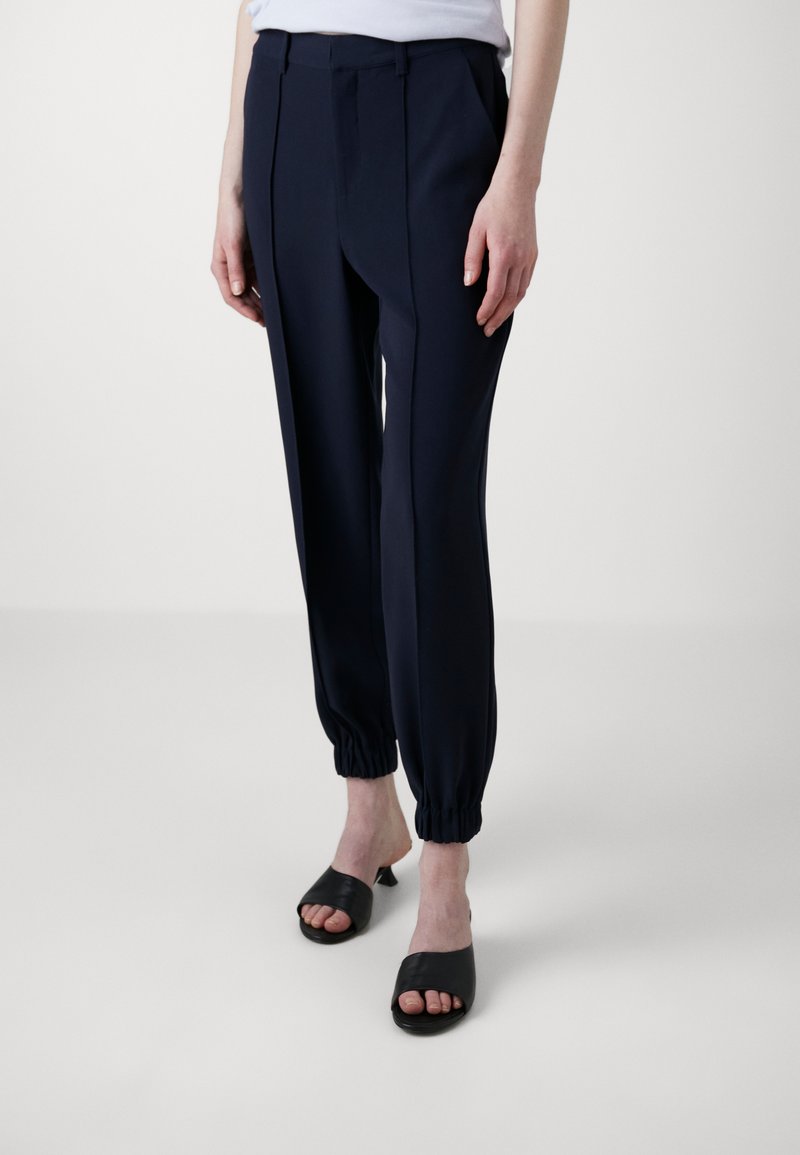 Marineblauwe tapered broeken met elastische boorden, met een gladde textuur en geplooide voorkant. Gecombineerd met zwarte openstaande muiltjes.