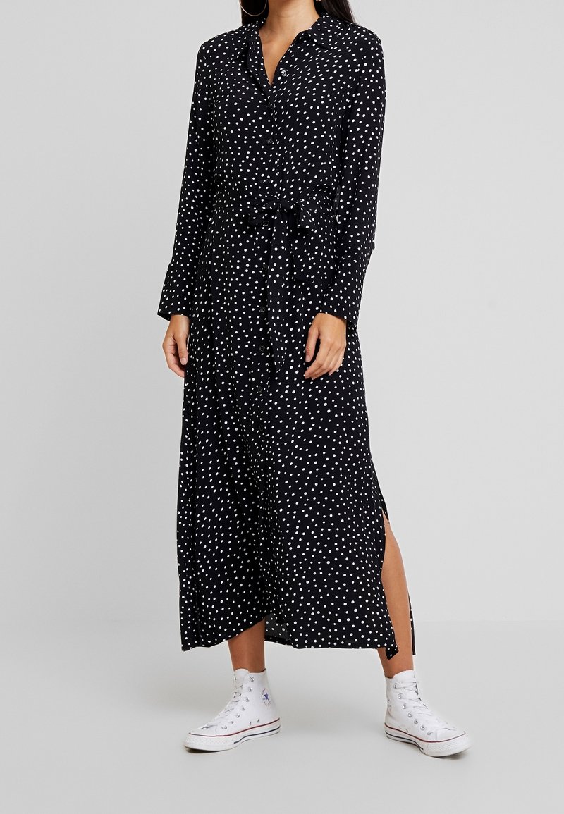 Robe longue noire à pois blancs, avec un devant boutonné, taille cintrée par un lien et fentes latérales. Associée à des baskets montantes blanches.