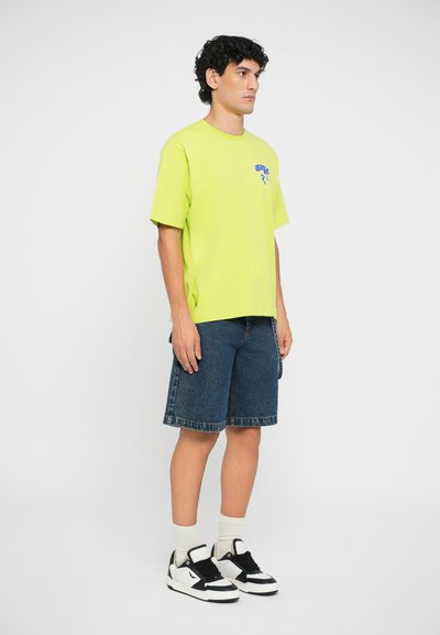 Neon green t-shirt with a blue logo on the chest, paired with denim shorts. Footwear includes black and white sneakers with cream socks.