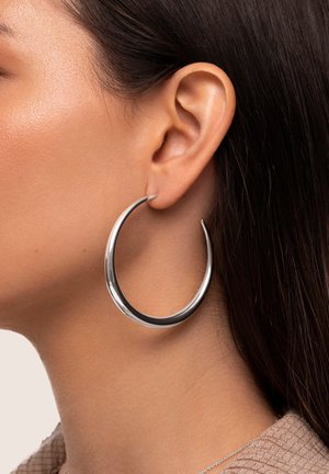Gran pendiente de aro plateado grande con una superficie pulida y reflectante, que presenta un diseño circular y una textura suave, llevado en la oreja.