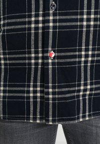 Camicia a quadri blu navy e crema in cotone con colletto button-down, caratterizzata da un bottone accentato rosso nella parte inferiore.