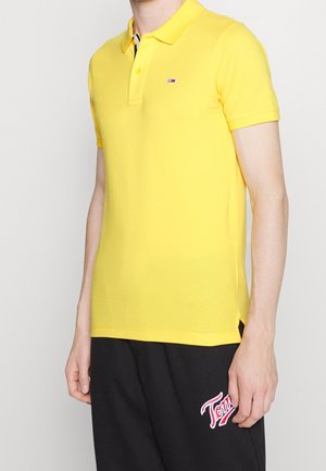 Žluté polo tričko s límečkem, krátkými rukávy a bočními průstřihy. Má malý logo na hrudi a hladkou bavlněnou texturu.