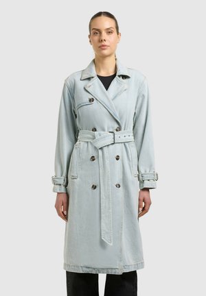 Frau mit zurückgebundenem Haar, die einen hellen Trenchcoat aus Denim mit Gürtel über schwarzer Hose trägt und vor einem einfarbigen grauen Hintergrund steht.