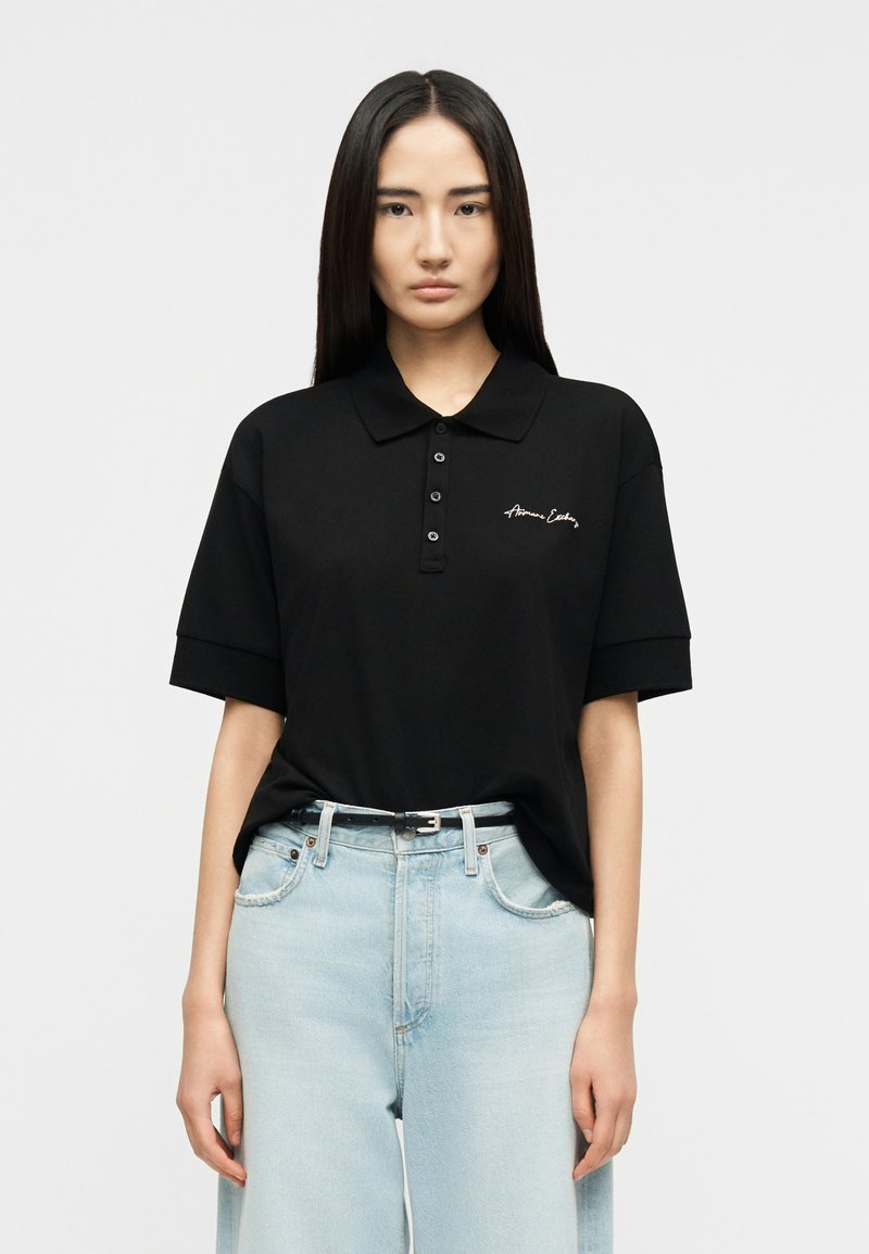 Γυναίκα με μακριά ίσια μαλλιά, ντυμένη με μαύρο πόλο με κοντά μανίκια και λογότυπο, και ανοιχτό μπλε ψηλόμεσο τζιν, μπροστά σε απλό γκρι φόντο.