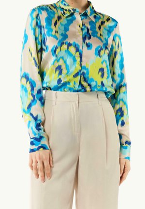 Femme portant un chemisier à manches longues boutonné avec un imprimé abstrait bleu, jaune et marine, rentré dans un pantalon beige taille haute.