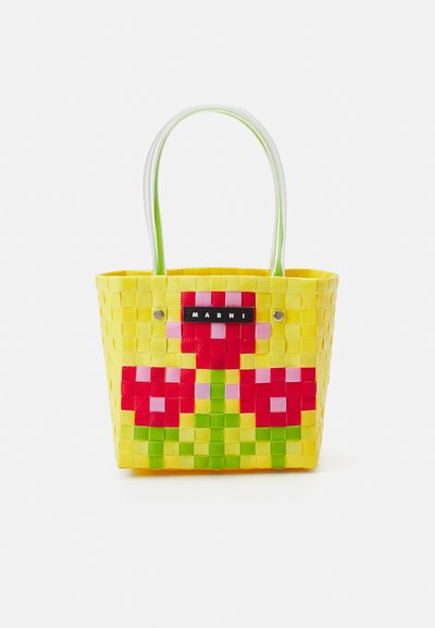 Saco de palha amarelo com padrão floral em vermelho, rosa e verde. Apresenta duas alças brancas e uma placa de logo preta.