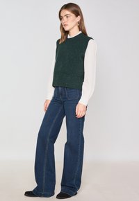 Maglione verde lavorato a maglia senza maniche con orlo a coste, abbinato a pantaloni a vita alta a gamba larga blu scuro e scarpe nere basse.