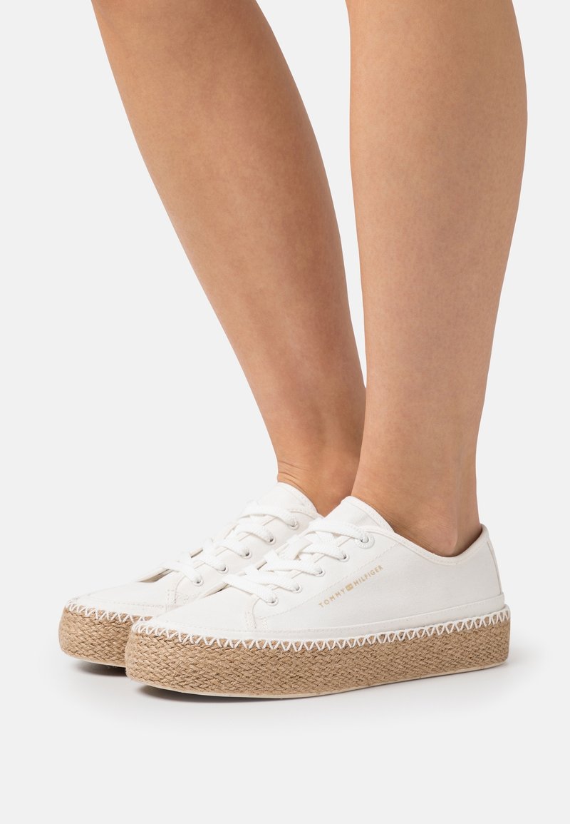 Jambes portant des baskets en toile blanche avec des semelles compensées en jute tissé et le logo "Tommy Hilfiger" sur le côté sur un fond blanc.