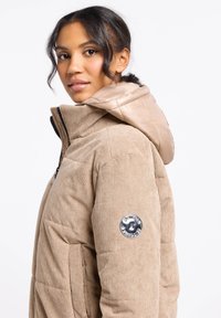 Karmelowa kurtka puchowa z sztruksu z błyszczącym beżowym kapturem, zamek błyskawiczny oraz okrągła naszywka z logo na lewym rękawie. Teksturowany materiał z pikowanym wzorem.