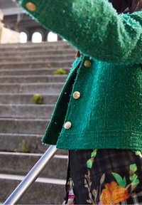 Veste verte texturée avec des boutons dorés, coupe courte. Tissu à motifs floraux visible en dessous et rampe métallique en arrière-plan.