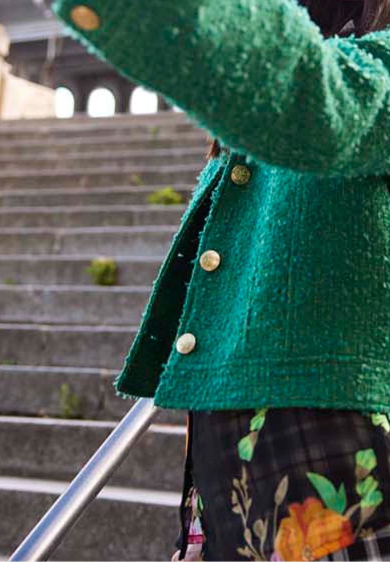 Veste verte texturée avec des boutons dorés, coupe courte. Tissu à motifs floraux visible en dessous et rampe métallique en arrière-plan.
