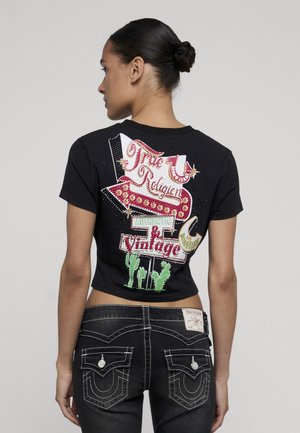 Donna con capelli scuri raccolti in uno chignon, indossa un crop top nero con grafiche colorate in stile vintage e jeans scuri con cuciture bianche.