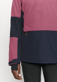 Person iført en tofarvet jakke i pink og marineblå med lynlåsdetalje og sorte bukser, der viser venstre arm og delvist torso.