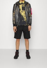 Svart huva jacka med reflexdetaljer, i kombination med en skjorta med gult och svart mönster samt svarta shorts. Avslutat med svarta kängor.