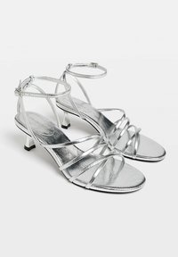 Sandales argentées à lanières avec une finition métallique brillante, dotées d'un petit talon et d'une bride de cheville, conçues avec plusieurs fines lanières à l'avant.
