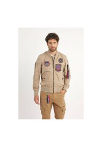 Veste bomber beige avec des patchs, fermeture éclair, poignets et ourlet côtelés, et détail de porte-clés avec des accents rouge, blanc et bleu.