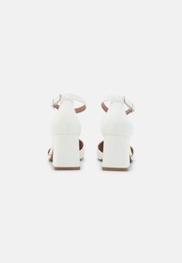 Sandales à talons blancs avec un talon carré, une sangle à la cheville et une texture lisse semblable au cuir. Design à dos ouvert avec une doublure intérieure marron contrastante.