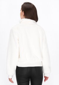 Chaqueta de forro polar blanco con cuello alto, puños acanalados y dobladillo. Textura suave y ajuste relajado. Combinada con pantalones negros de corte slim.
