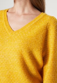 Femme portant un pull jaune texturé à col en V et manches longues, avec la clavicule visible et des cheveux bruns reposant sur l'épaule.
