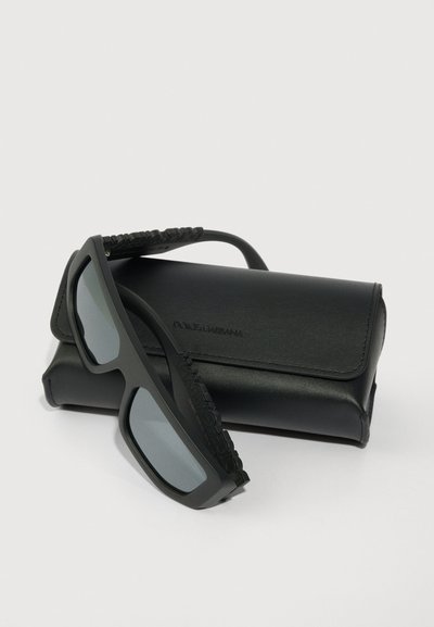 Melnas taisnstūra saulesbrilles ar gaiši pelēkiem stikliem, kas atpūšas uz melna ādas Dolce & Gabbana zīmola futrāļa.