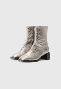 Botines metálicos plateados con punta afilada, que cuentan con una cremallera lateral, tacón bajo y una textura suave y reflectante.