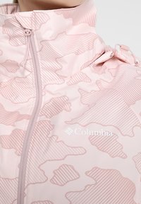 Veste rose clair avec capuche, motif camouflage abstrait, fermeture éclair frontale et logo Columbia sur la poitrine.