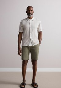 Kortærmet, lys beige strikbluse med knaplukning og struktureret mønster, parret med lysegrønne shorts og mørkebrune sandaler.
