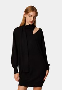 Vestito nero in maglia con collo alto, maniche lunghe e un taglio laterale. Presenta un lungo dettaglio simile a una sciarpa che drappeggia dal collo.