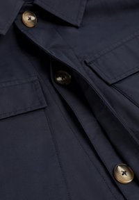 Camisa abotonada azul marino con dos bolsillos en el pecho, con grandes botones marrones y una textura de tela elegante. Cuello doblado y detalles de costuras limpias.