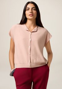 Vrouw met lang, donker haar, gekleed in een lichtroze, korte mouwen blouse met knopen en donkere rode broek, met één hand in de zak en een armband om de pols.