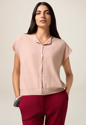 Donna con capelli lunghi e scuri, che indossa una camicetta rosa chiaro a maniche corte con bottoni e pantaloni rosso scuro, con una mano in tasca e un braccialetto al polso.