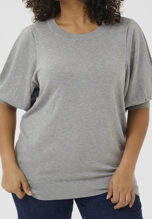 Femme portant un pull tricoté gris clair à manches courtes bouffantes et un jean bleu foncé, debout devant un fond uni.