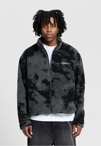 Giacca in poliestere nera e grigia con colletto alto, chiusura con zip e motivo camouflage. Presenta un logo sul lato sinistro del petto e polsini a costine.