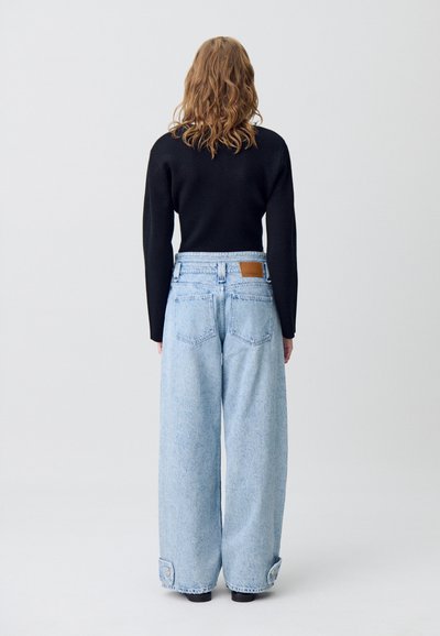 Jeans larghi di colore blu chiaro con vita alta, leggermente sbiaditi e orli risvoltati. Una patch in pelle marrone è visibile sulla parte posteriore della vita.